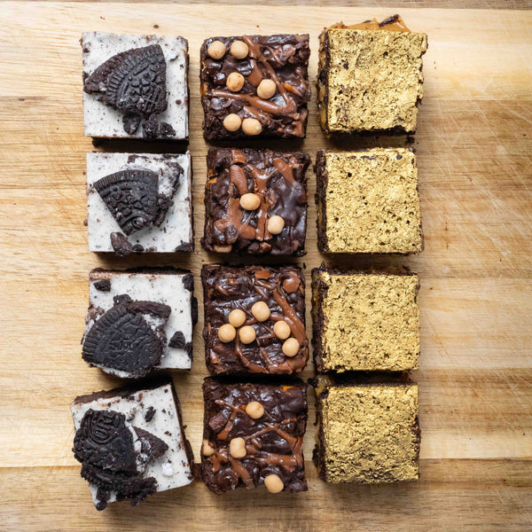 Twelve square brownies arranged in a 3-by-4 grid on a wooden cutting board. Each column is a different flavour of chocolate brownie