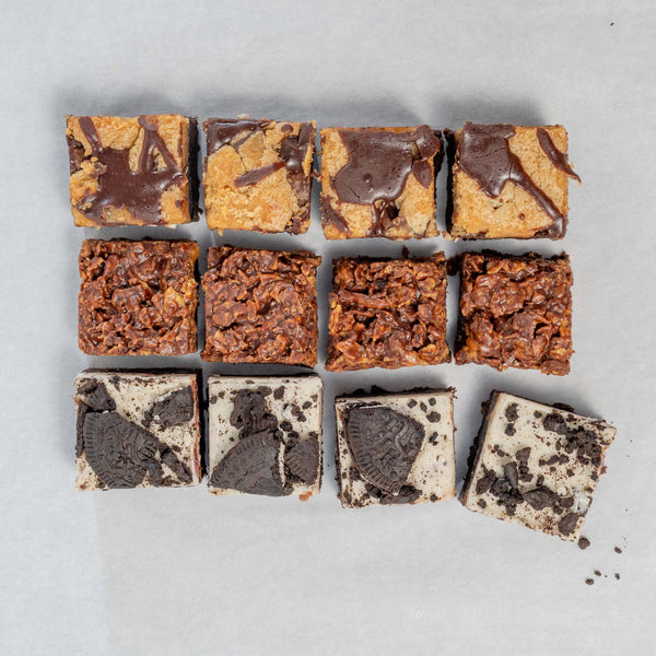 A box of assorted brownies with different flavours like cornflake biscoff, cookie dough and cookies and cream, arranged in a grid pattern on a grey background.