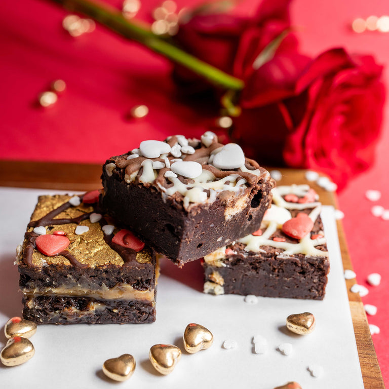 Chocolate brownies with gold and red decorations on a white surface with red roses in the background.