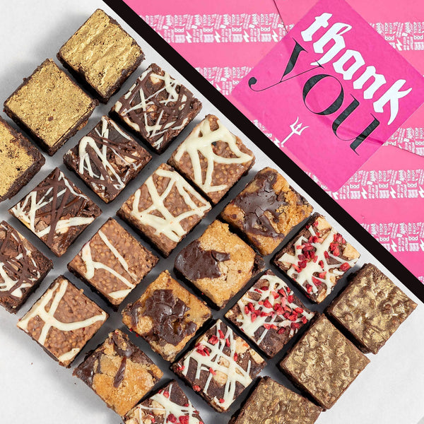 The Premier 24 Bite Brownie Box in Thank You gift wrap. Assorted brownies on a white surface with a pink 'thank you' giftwrap in the background.