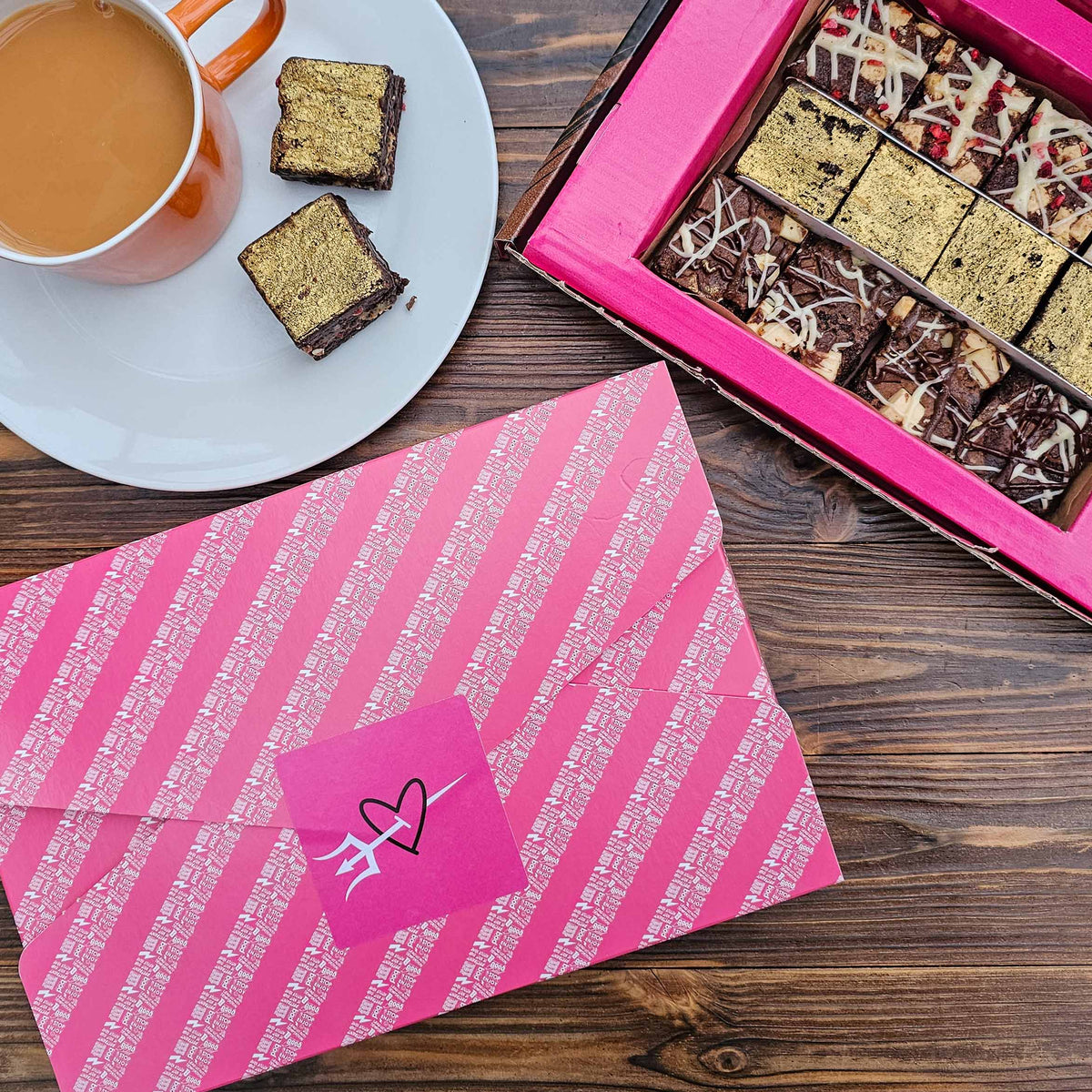 Pink striped gift wrap with a heart design, box of assorted brownies, and cup of coffee on a wooden surface.