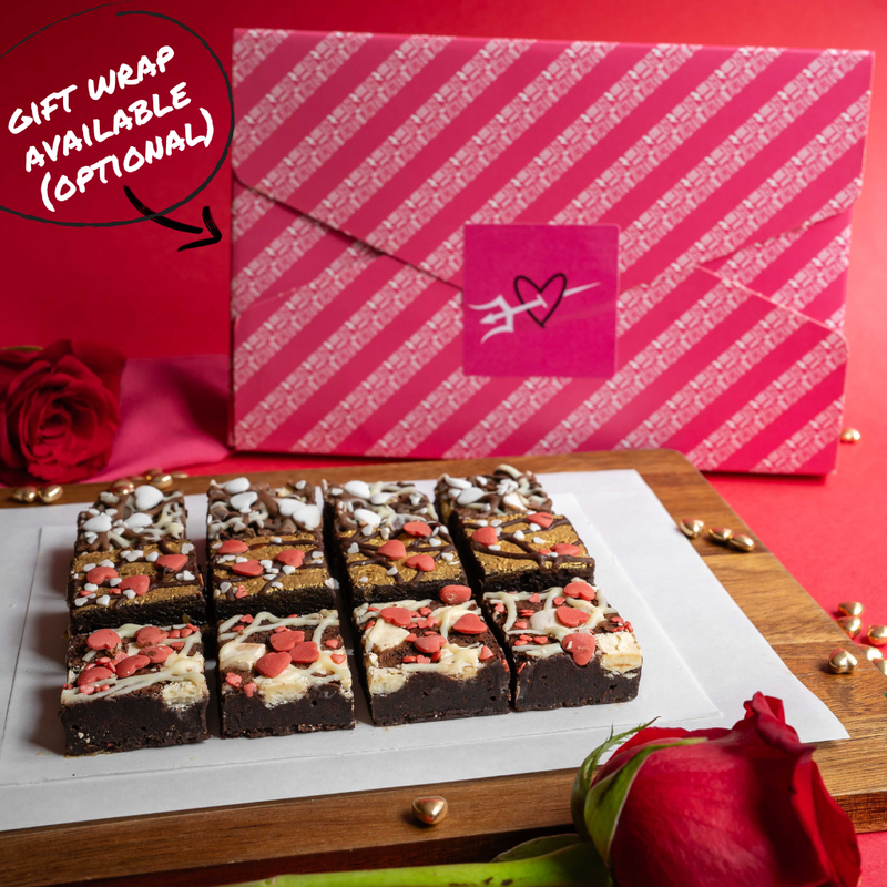 Brownie squares with Valentine's Day decorations on a wooden board, with a pink gift box and red roses in the background.