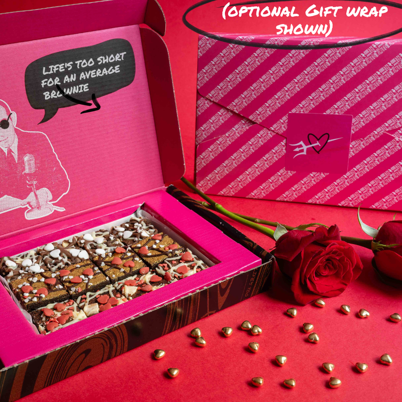 Open pink box of Bad Brownie chocolate brownies with a rose, gold candies and a gift wrapped Valentines Day box on a red background.
