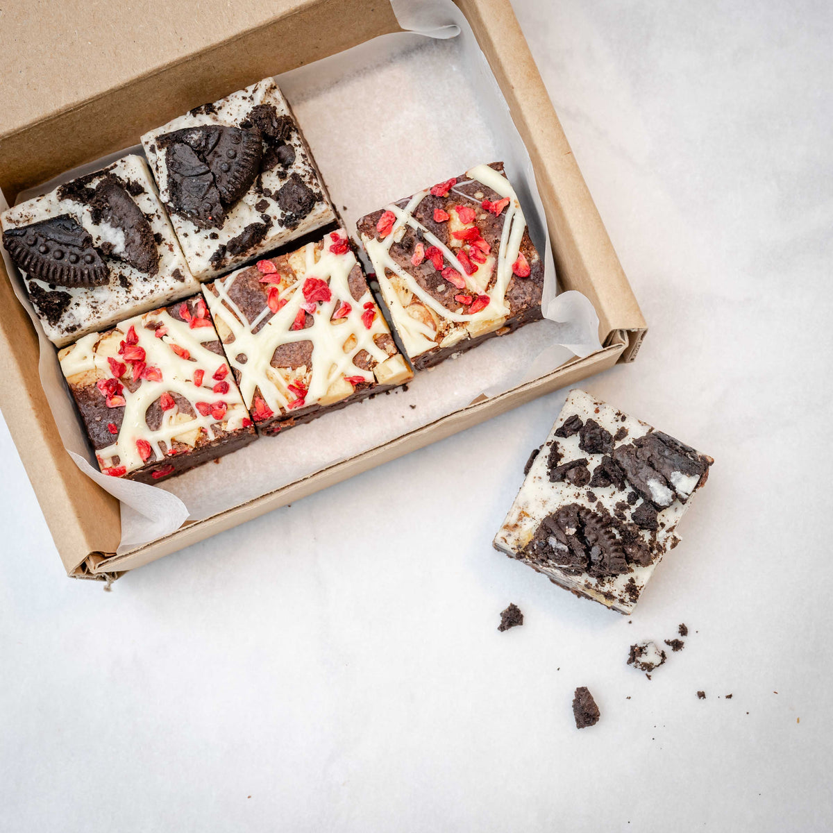 An array of six brownie bites from the Discovery 6 Bite Brownie Box, showcasing different flavors with visible chunks of raspberry and cookies. 5 bites are inside the box and one is outside