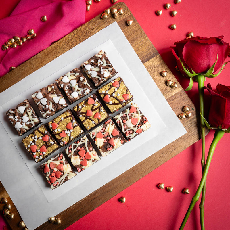 Romantic Valentines Day mixed flavour brownie bites which are covered in edible heart toppings, on a wooden board against a red background with gold hearts and red roses in the background 