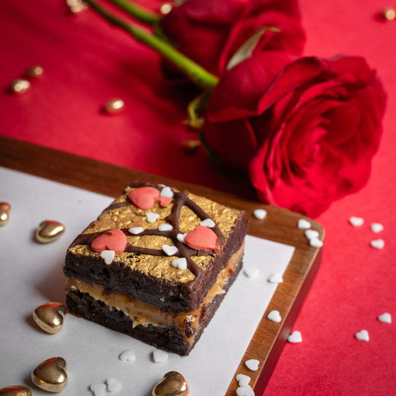 Gold-leaf topped salted caramel brownie with heart decorations on a red surface with roses