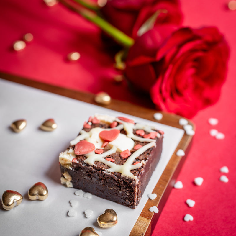 Raspberry and white chocolate brownie with white chocolate and red heart toppings on a red background with roses.