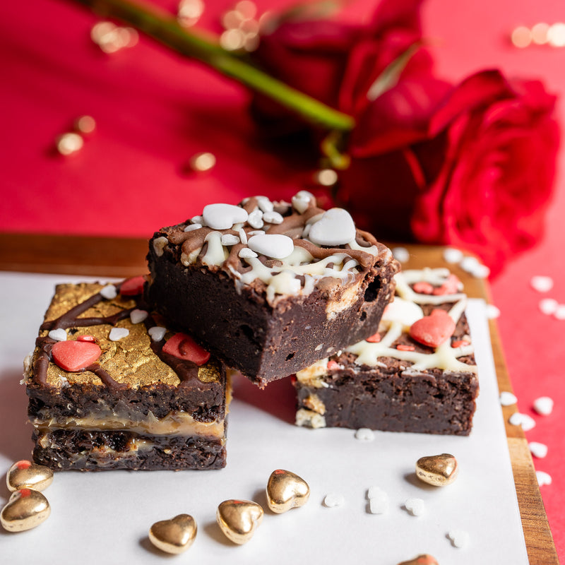 Chocolate brownies with white chocolate chips and red heart-shaped candies on a wooden board with red roses in the background.