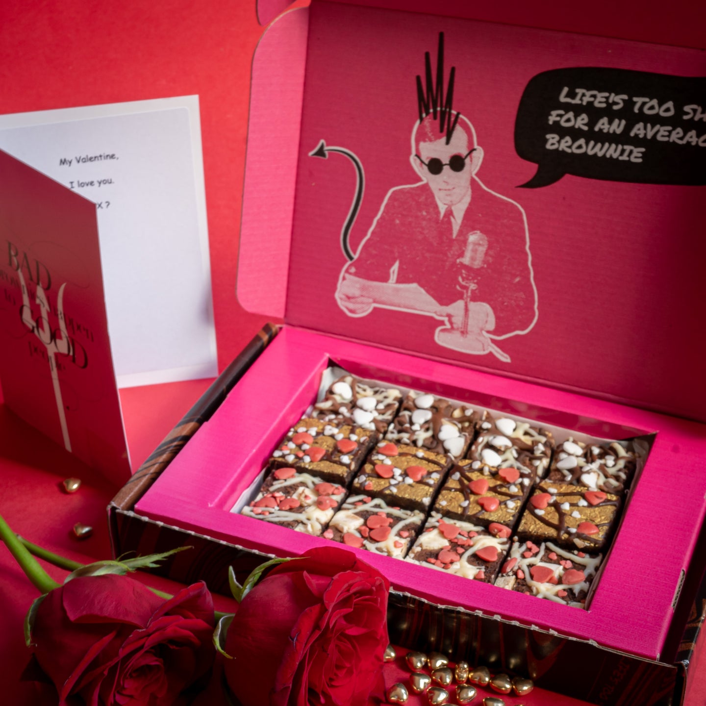 Open pink Valentine's chocolate brownie postal delivery box with assorted Brownies by Bad Brownie, a card, and red roses on a red background.