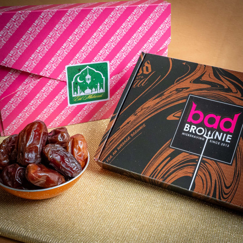 Box of Bad Brownie with a bowl of dates on a textured surface and Eid gift wrapped present in the background