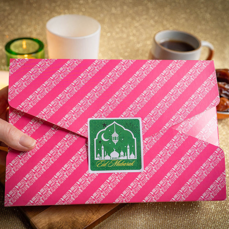 Pink Eid Mubarak brownie gift box with white text and a green logo held by a hand, with a cup of coffee and dates in the background.