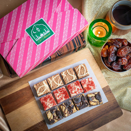 Assorted chocolate brownies on a wooden board with a pink Eid gift box, dates, and a candle in the background.