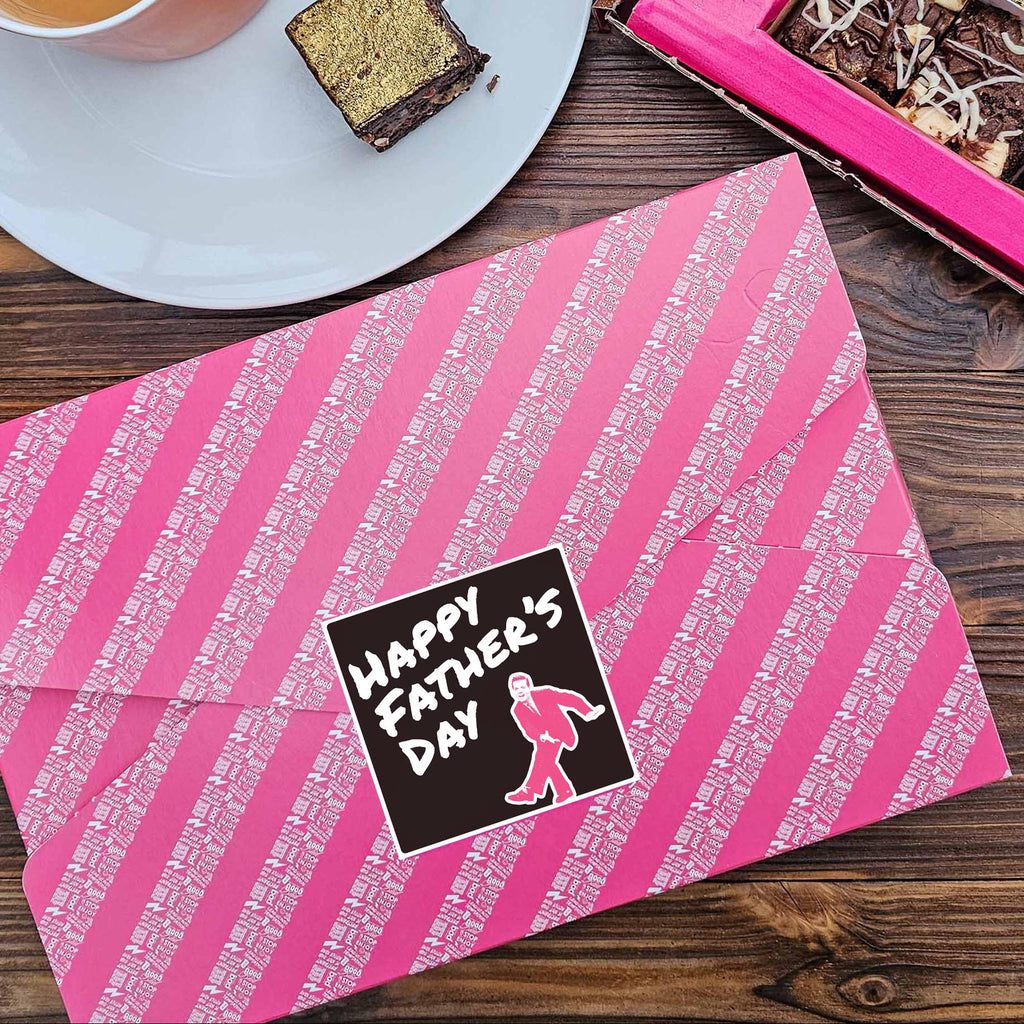 Pink gift wrapped box of brownies with a black Happy Father's Day Sticker, on a wooden tabletop with open brownies and a cup of tea