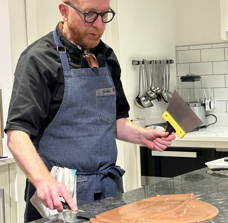 Paul A Young in a kitchen wearing an apron, tempering chocolate on a marble counter.