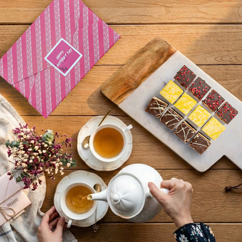 tabletop showing flavoured brownies on marble platter along with gift wrapped box and a lady's hands pouring tea into a cup next to a vase of flowers