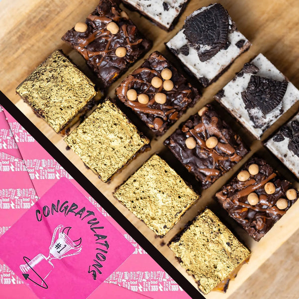 LIMITED EDITION Cosy Cravings Brownie Box in Congratulations gift wrap. An overhead view of assorted chocolate brownies arranged in a wooden tray, with bright pink congratulations gift wrap placed diagonally across the lower left corner.