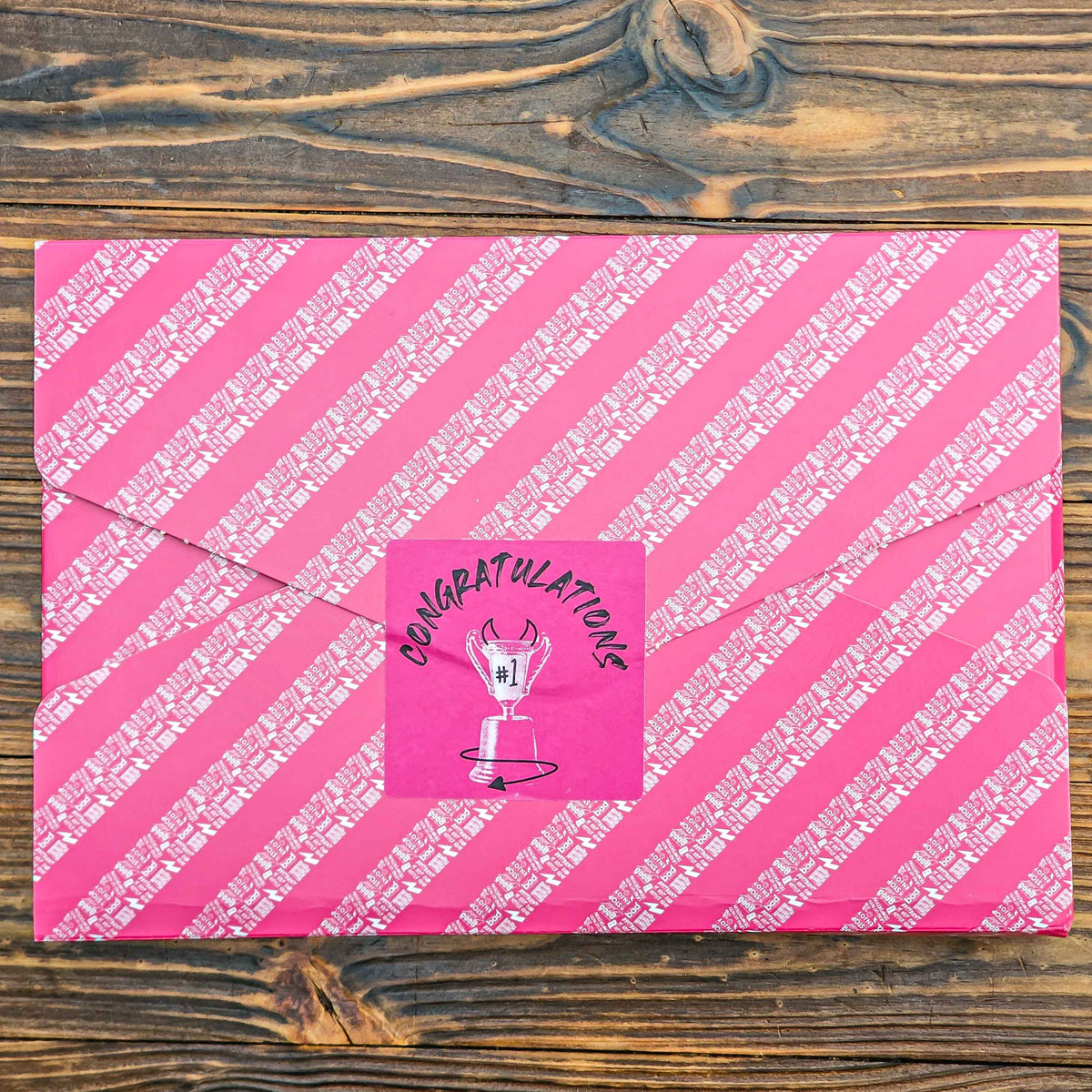 Pink gift wrapped brownie box with white diagonal stripes and a 'Congratulations' sticker on a wooden surface