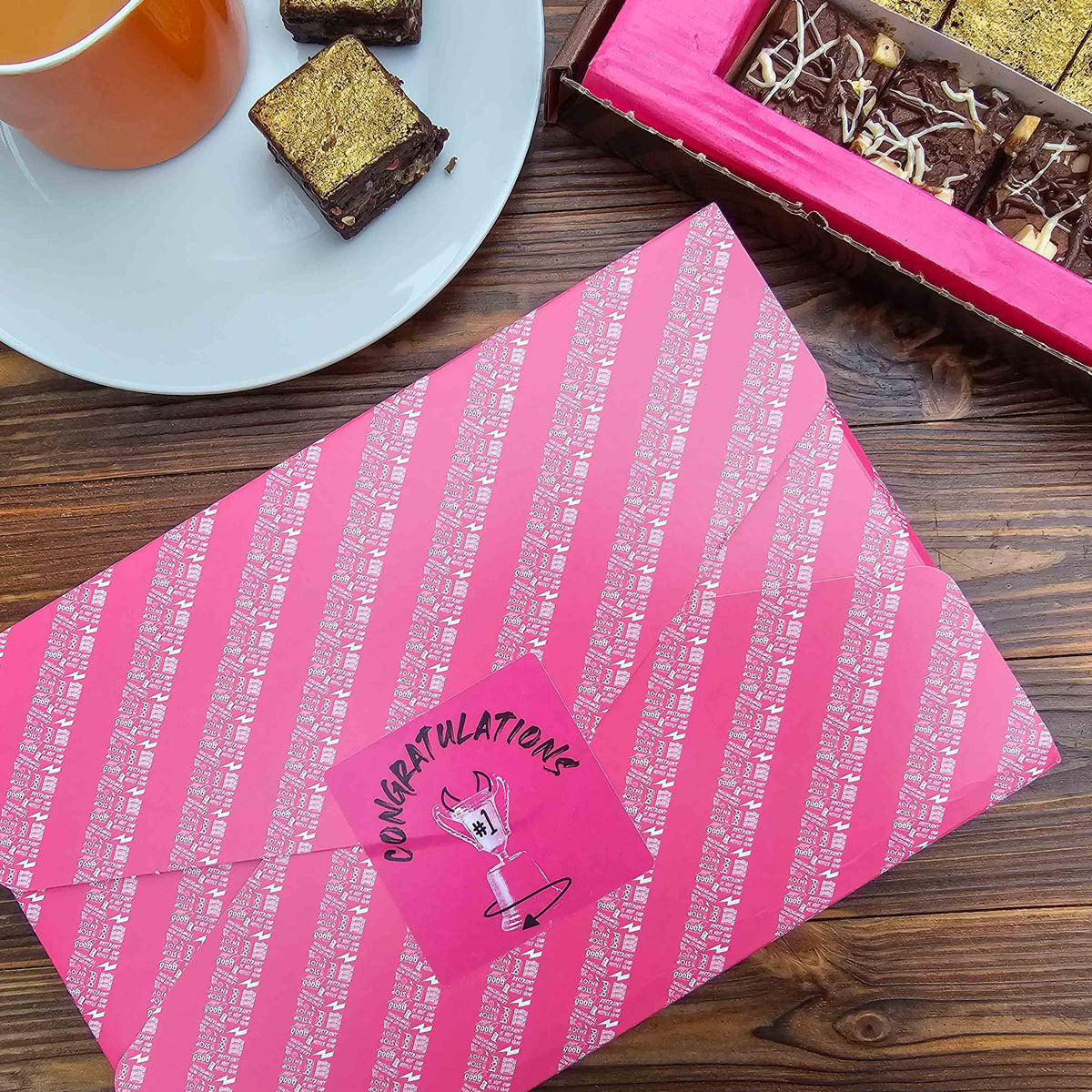 Pink gift wrapped brownie box with 'Congratulations' on a wooden surface with chocolate brownies and a cup.