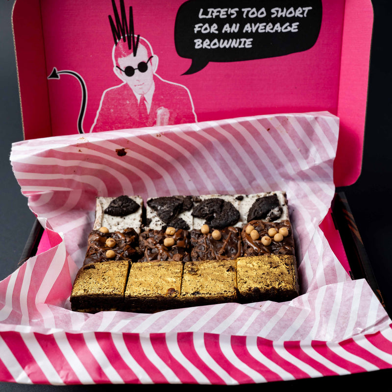 A boxed assortment of brownies presented in a pink and white striped box with a bright pink lid featuring a cartoon illustration and the text 'LIFE'S TOO SHORT FOR AN AVERAGE BROWNIE.' The brownies are arranged in rows 