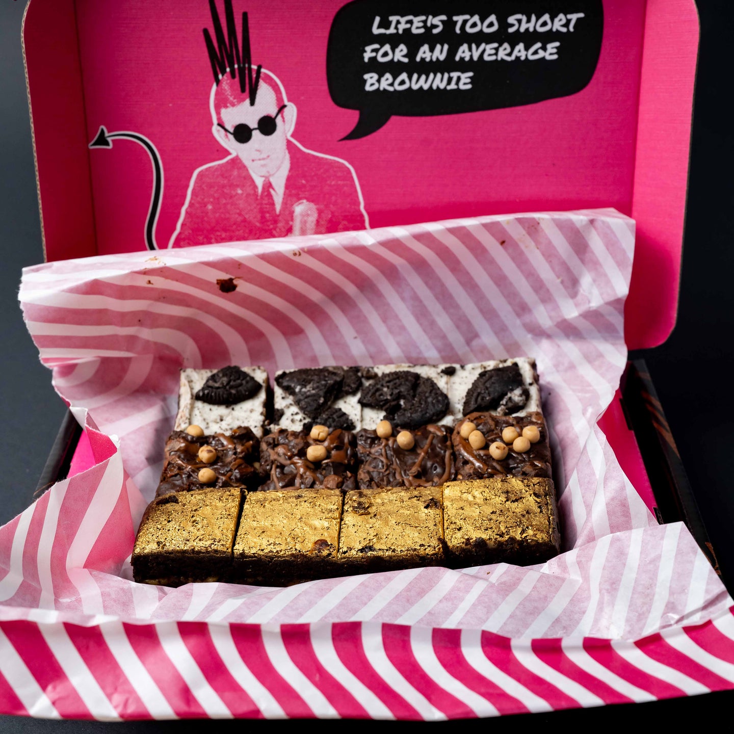 A boxed assortment of brownies presented in a pink and white striped box with a bright pink lid featuring a cartoon illustration and the text 'LIFE'S TOO SHORT FOR AN AVERAGE BROWNIE.' The brownies are arranged in rows 