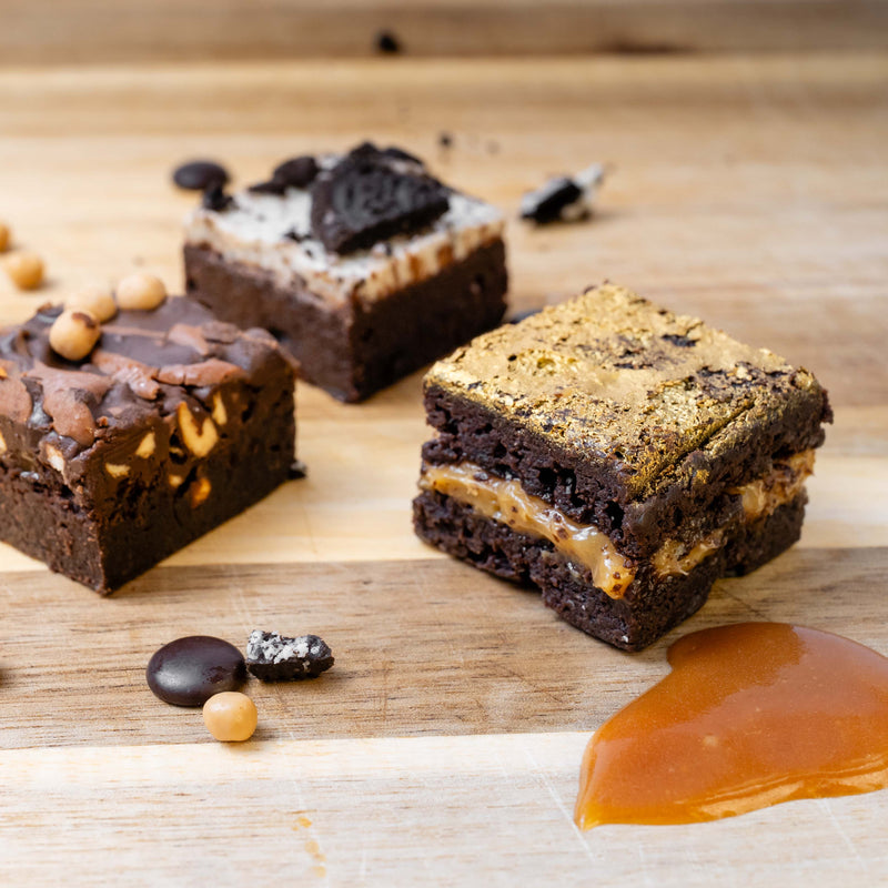 Three flavoured brownies on a wooden board with caramel sauce, choc chips, and cookie crumbs. The front brownie shows a salted caramel layer, while the others feature chocolate chips, pretzels, and cookie pieces.