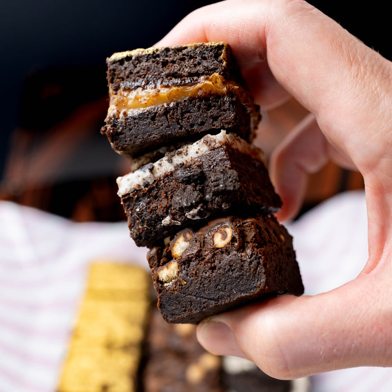 A hand holding a stack of three gourmet chocolate brownies with caramel, cookies and cream, and pretzels, set against a blurred background of more brownies.