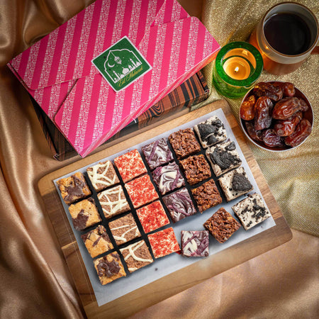 Assorted chocolate brownies on a wooden board with a pink Eid gift box, dates, and a candle in the background.