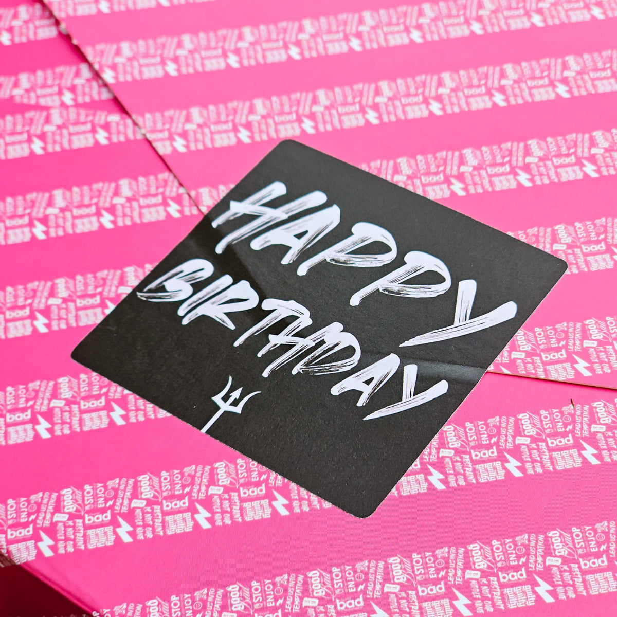 A close-up of a a pink gift wrapped box of Birthday brownies with text pattern and a black sticker that reads 'Happy Birthday' in white font.