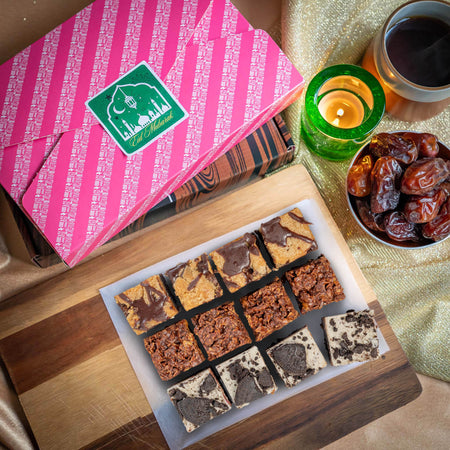 Assorted chocolate brownies on a wooden board with a pink Eid gift box, dates, and a candle in the background.
