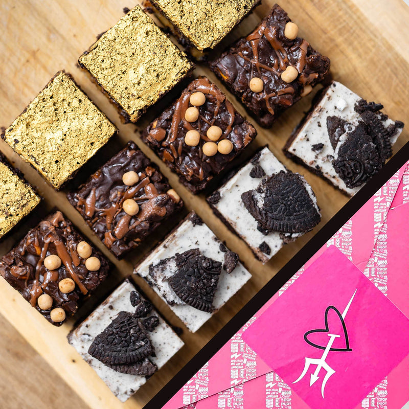 A top-down view of assorted chocolate brownies arranged on a wooden board, with a bright pink branded gift wrap partially visible in the lower right corner.