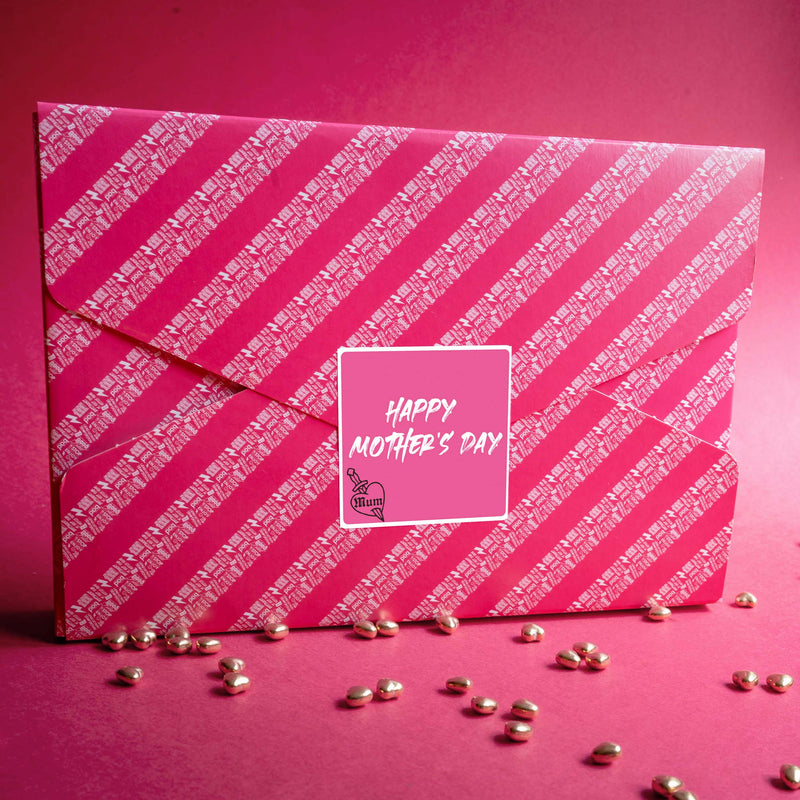 pink gift wrapped bad brownie postal box on a pink background with Happy Mother's Day sticker in the middle of the box with metallic sprinkles at the base