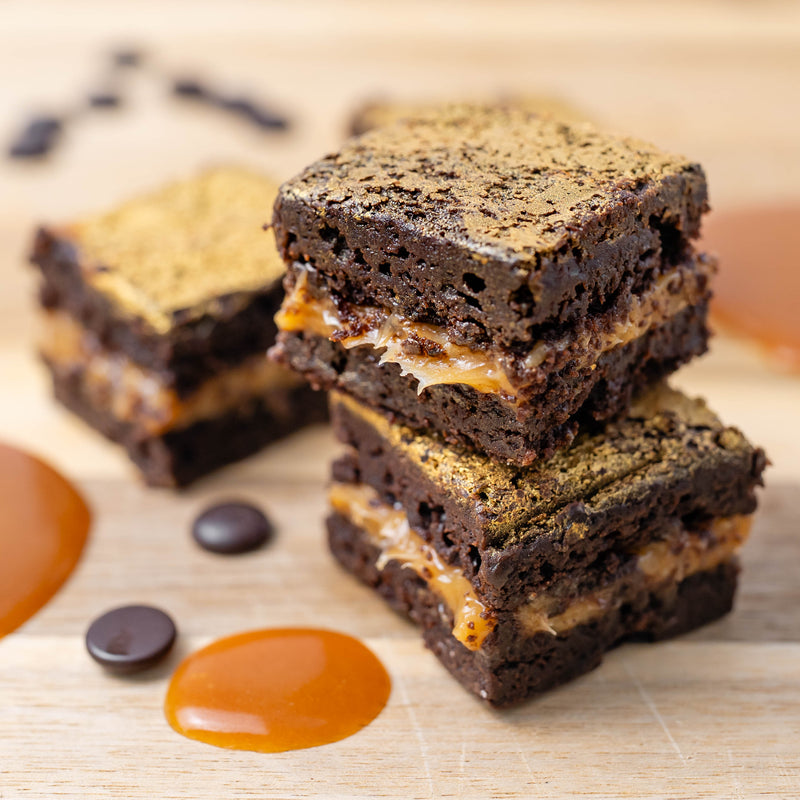 Three stacked chocolate brownies with salted caramel filling on a light wooden board, with scattered chocolate chips and caramel drizzle around them.