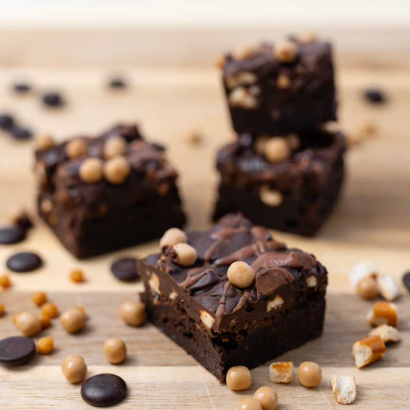Four pretzel chocolate brownies topped with chocolate and crispy caramel balls, arranged on a light wooden board with scattered ingredients.