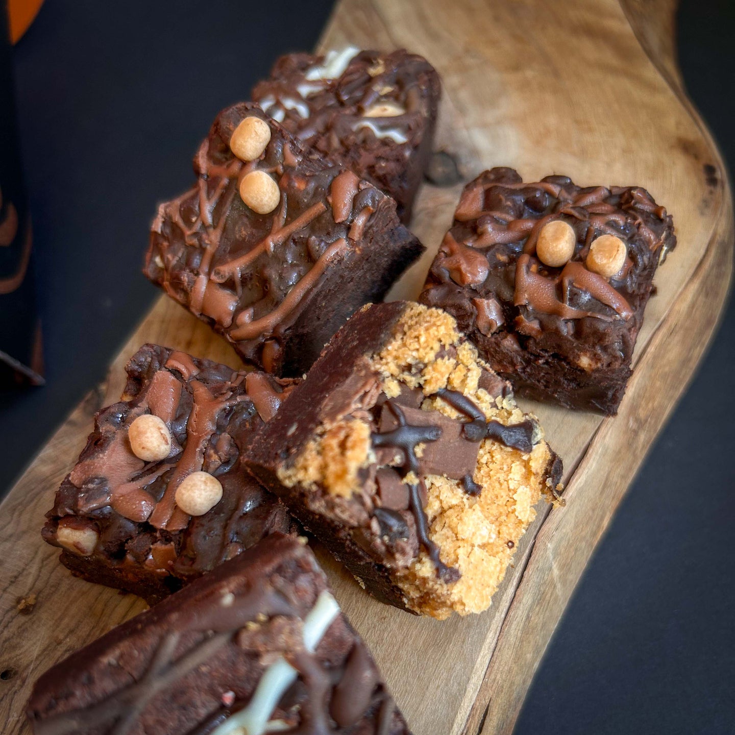 Six chocolate brownies arranged on a rustic wooden serving board, decorated with drizzles of chocolate, cookie dough, and small caramel balls, photographed against a dark background.