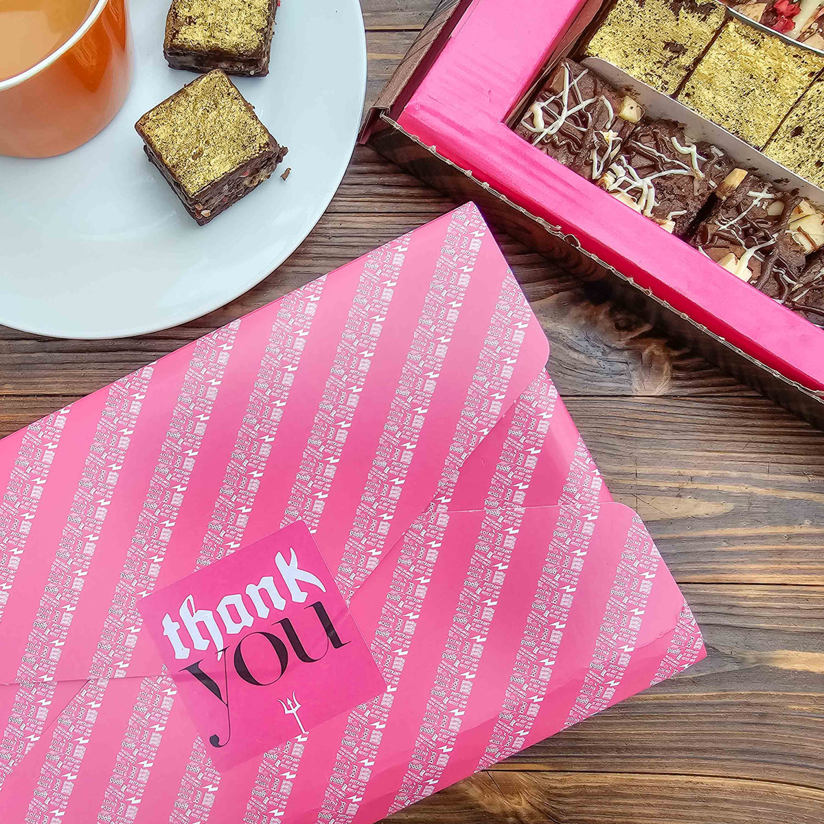 Pink gift wrapped box with 'thank you' text on a wooden surface with chocolate brownies and a cup.