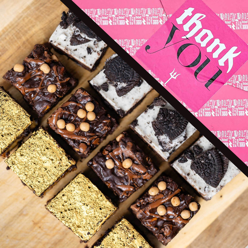 An assortment of gourmet chocolate brownies arranged on a wooden board, with a bright pink gift wrap labelled 'thank you' placed diagonally across the top right.
