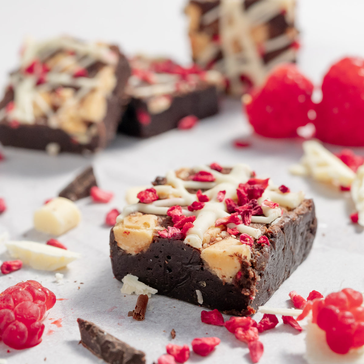 close up picture of a white chocolate and raspberry dark chocolate brownie with chocolate chips and raspberries on a neutral background